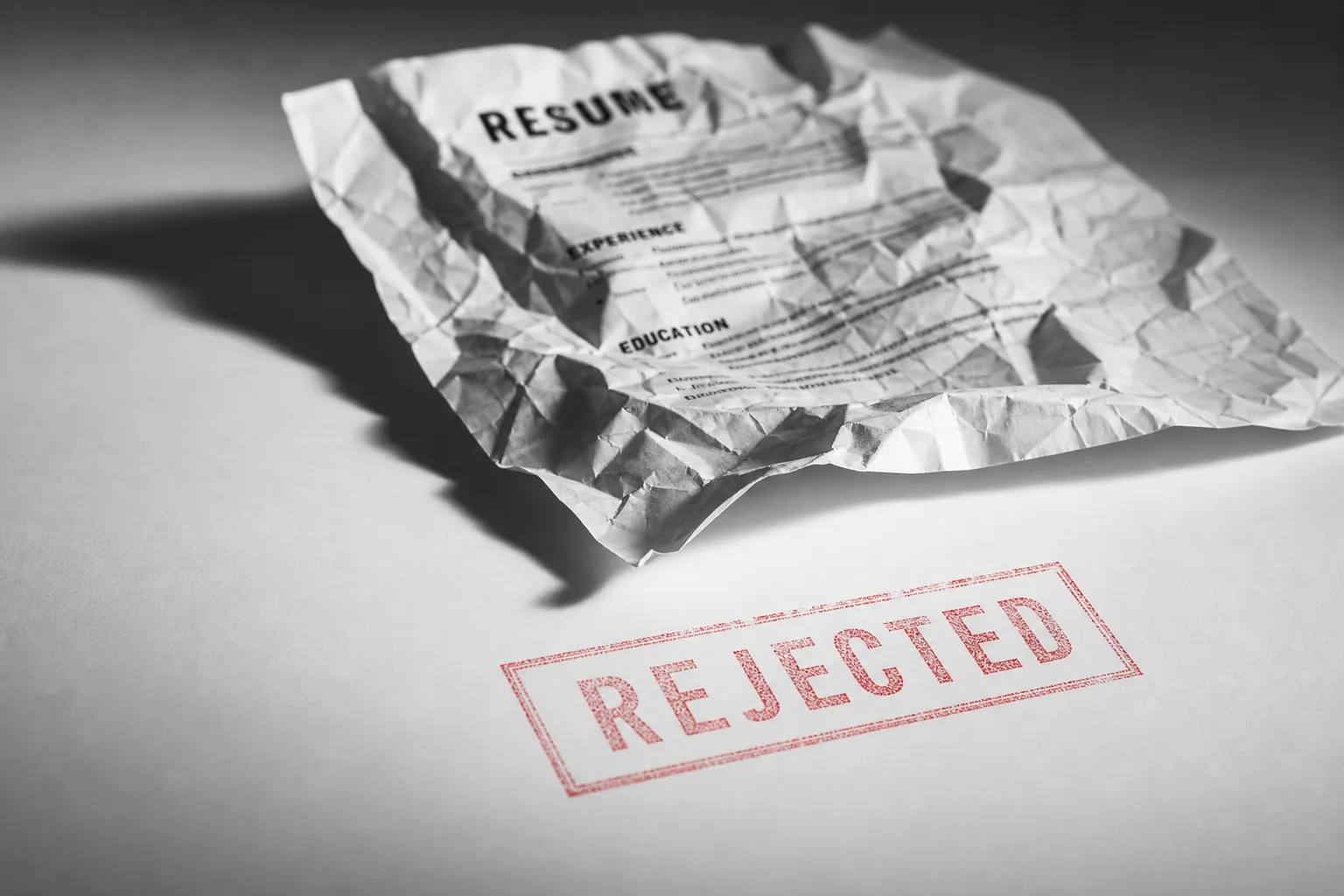 A single crumpled paper resume sitting alone on a stark white surface, dramatically underlit from one side, black and white editorial photography style. The resume is partially crushed, slightly out of focus in the background. In the sharp foreground, a faint red "REJECTED" stamp impression on the surface. Cold, clinical, bureaucratic feeling. No people. No faces. High contrast. The mood should feel systemic and impersonal, not emotional or melodramatic.